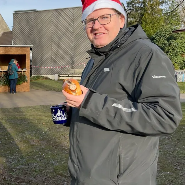 Pfarrer Fresia mit Bratwurst im Brötchen in der einen Hand und Glühwein in der anderen